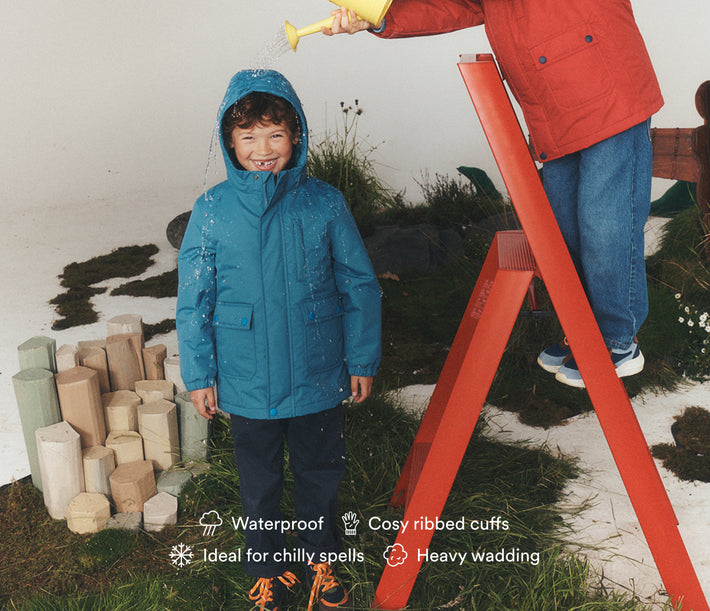 A child wearing a bright blue waterproof hooded raincoat stands on a grassy set while an adult in a red jacket pours water from a yellow watering can. The child's raincoat has 'KAVU' printed near the bottom hem.