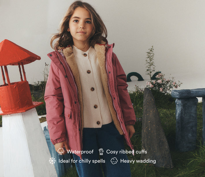 A young girl wearing a dusty pink children's parka with a beige sherpa-lined collar, standing in front of a white wall with decorative garden props. Text overlays at the bottom highlight product features: waterproof, cosy ribbed cuffs, ideal for chilly spells, and heavy wadding.