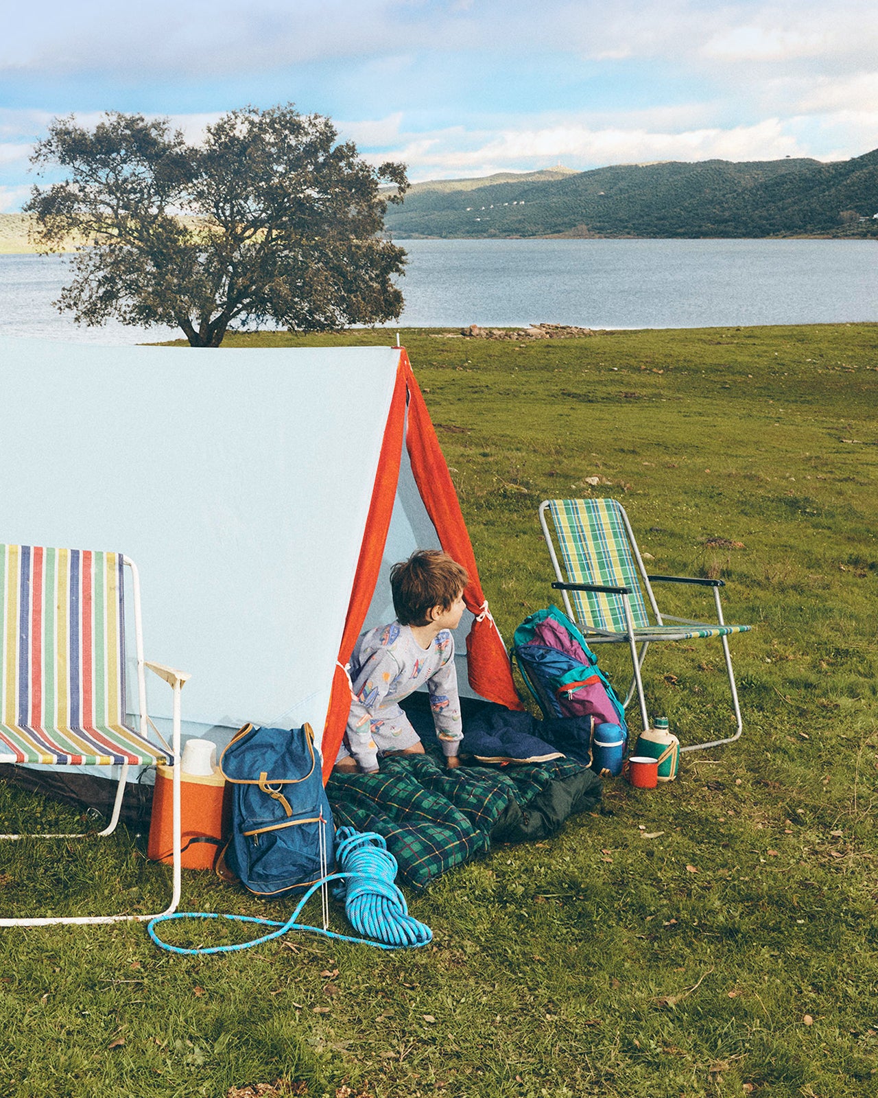Child playing under a small red tent in a grassy area by a lake.