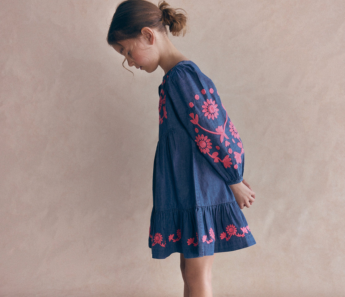 A young girl wearing a dark blue denim dress with pink floral embroidery, standing against a textured beige background.