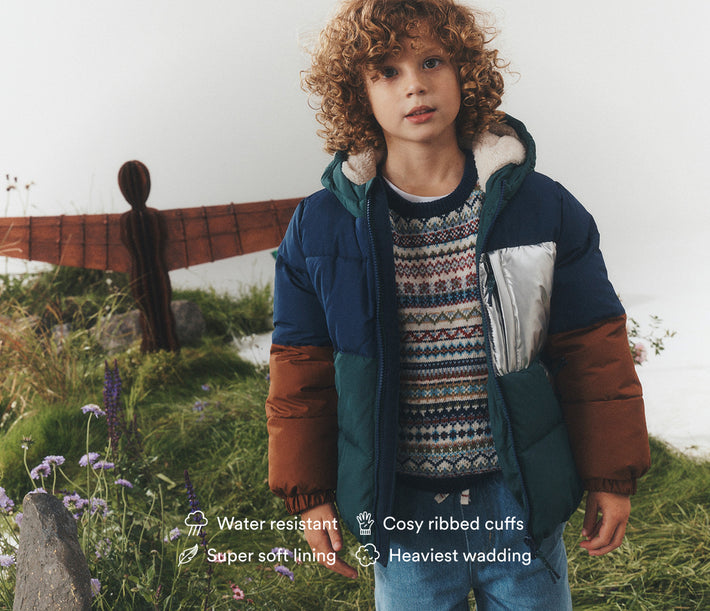 A child wearing a color-blocked puffer jacket outdoors on a grassy hill with a blurred angel sculpture in the background. The jacket is navy blue on the upper body, teal on the lower body, and brown on the sleeves, with a silver pocket and white faux-fur collar.