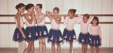 Group of young ballet dancers in a studio, wearing matching outfits with navy skirts and pink tops.