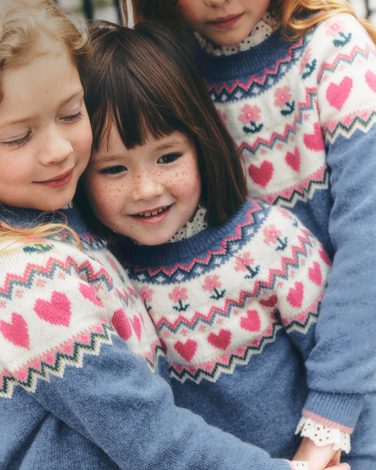 Three children wearing matching blue and cream knit sweaters with pink heart and floral Fair Isle patterns, hugging closely in a warm, close-up lifestyle shot.