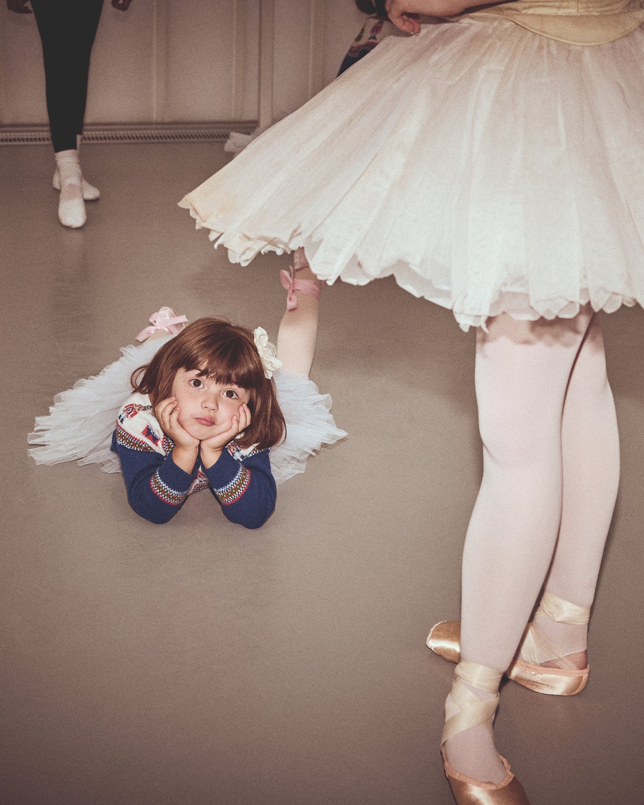 Child in a tutu lying on the floor with a ballet dancer standing nearby.