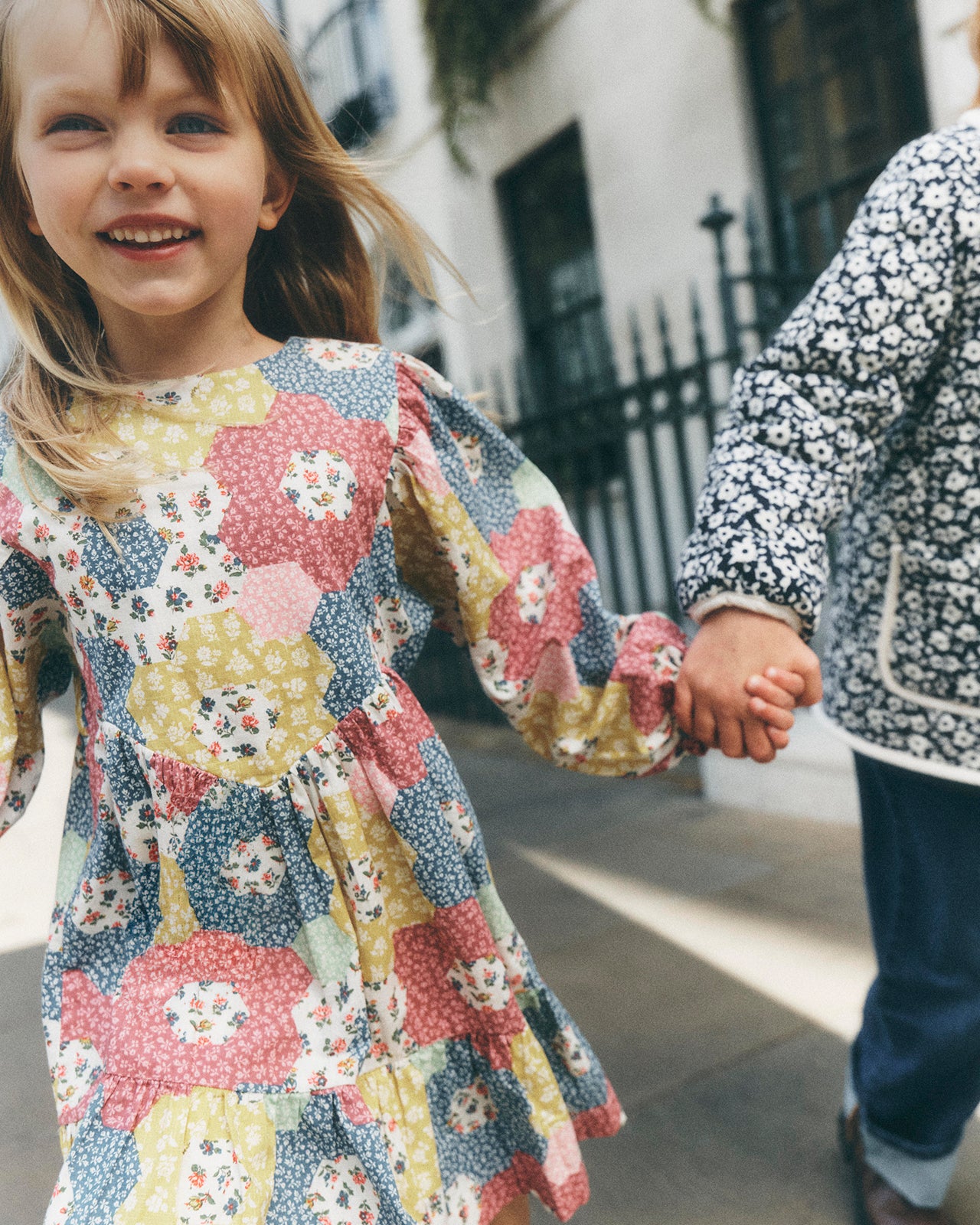 A young girl smiling and holding hands with another child outdoors. The girl wears a long-sleeve dress with a colorful floral and geometric patchwork pattern in pink, yellow, blue, and white.