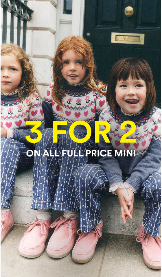 Three young girls sitting on stone steps in front of a dark door, wearing matching knit sweaters and trousers in pink, white, and navy. Large yellow text across the center reads '3 FOR 2' with smaller white text below reading 'ON ALL FULL PRICE MINI'.