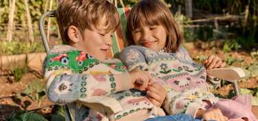Two children sitting on a bench in a garden, wearing patterned sweaters.