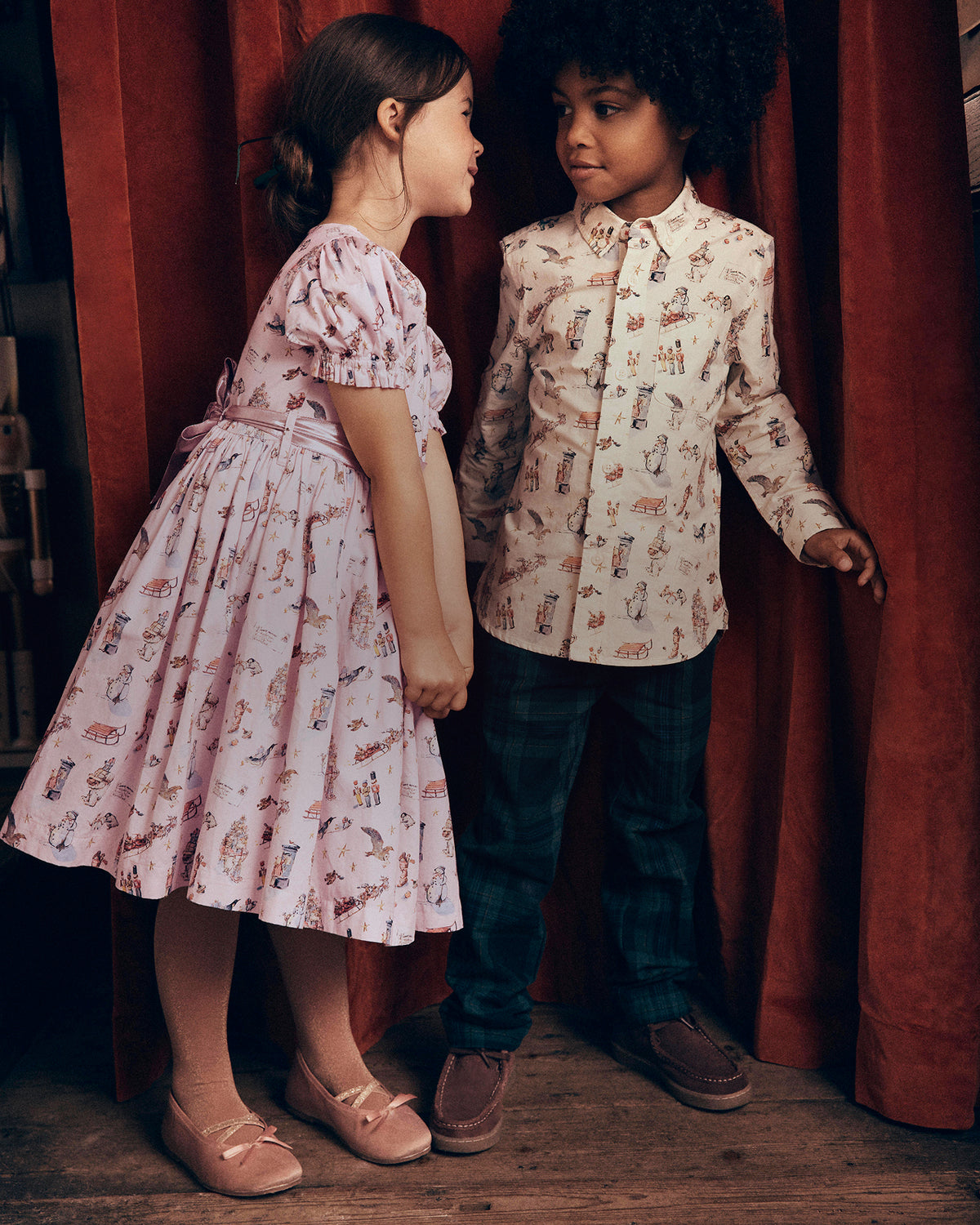 A young girl in a pink patterned dress and a young boy in a cream patterned button-up shirt and dark jeans, standing in front of a red curtain.