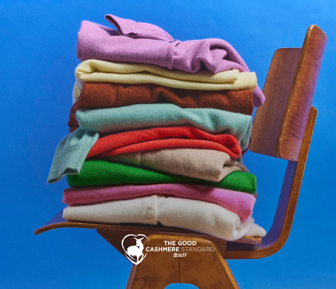 Stack of colorful cashmere sweaters on a wooden chair against a blue background, with 'The Good Cashmere Standard' logo.