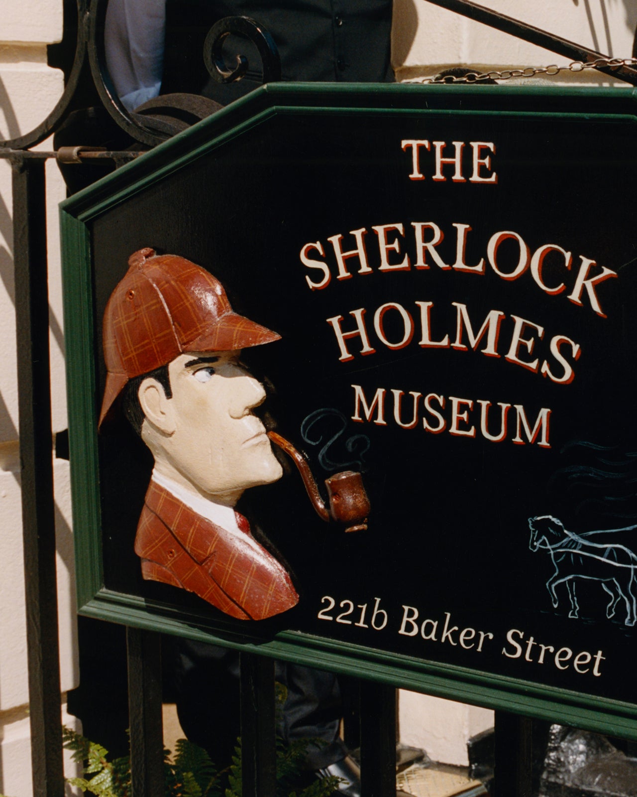 A close-up of a decorative sign for The Sherlock Holmes Museum, featuring a carved figure in a deerstalker hat and smoking a pipe, with white lettering on a dark green and black background.