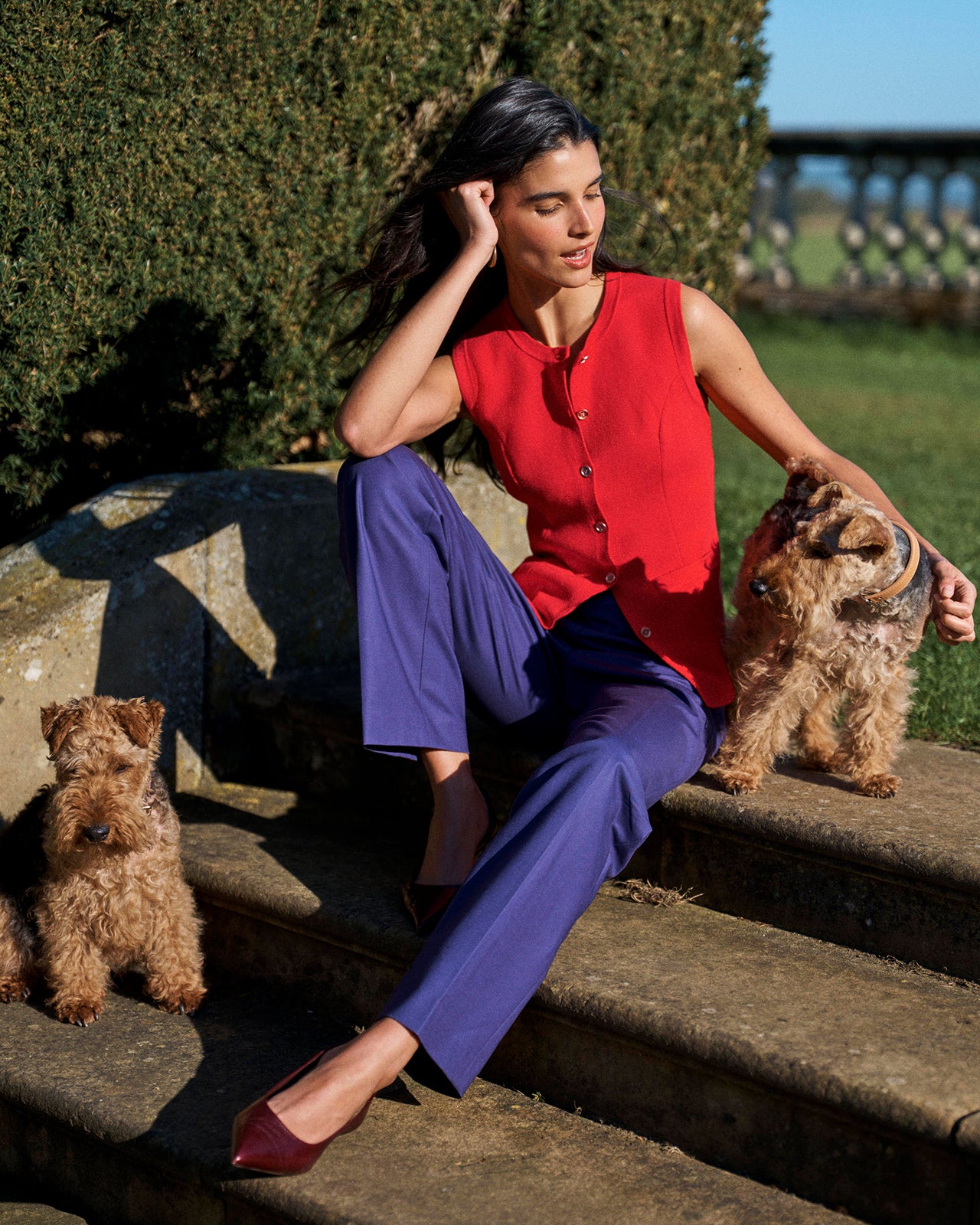 A woman sits on outdoor stone steps wearing a sleeveless bright red top and purple trousers, with two small dogs beside her in a sunny garden setting.