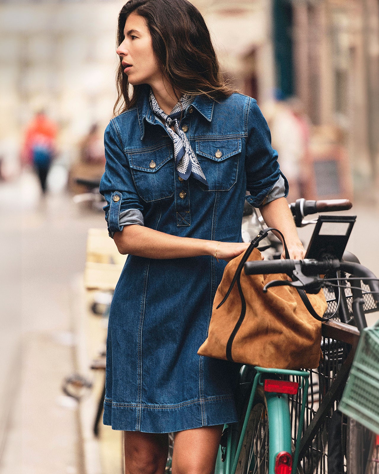 A woman wearing a blue denim shirt dress stands next to a bicycle in an outdoor street setting, holding a tan suede shoulder bag.