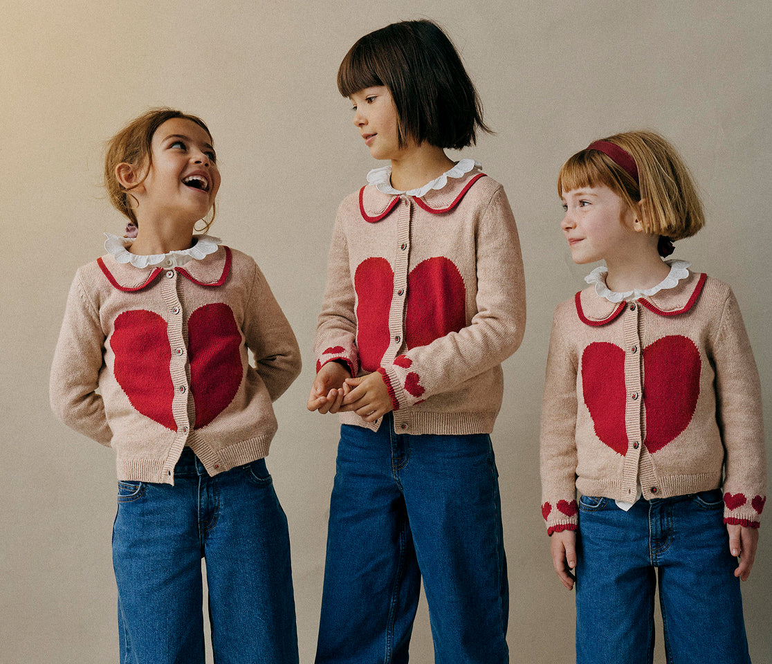 Three children wearing matching cardigans with red heart designs against a plain background