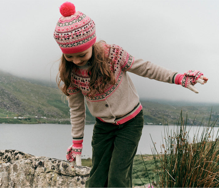 Person wearing a patterned sweater, hat, and gloves standing by a lake with mountains in the background.