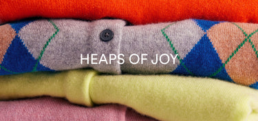 A close-up stack of folded knit sweaters in multiple colors, including orange, gray with blue and green argyle, yellow, and pink. White text across the center reads 'HEAPS OF JOY'.
