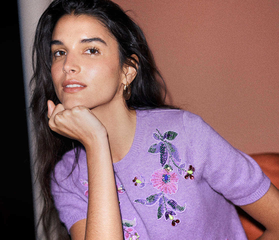 A woman with long dark hair rests her chin on her hand, wearing a lavender purple short-sleeve knit sweater with colorful floral embroidery and sequins on the front, set against a warm terracotta background.