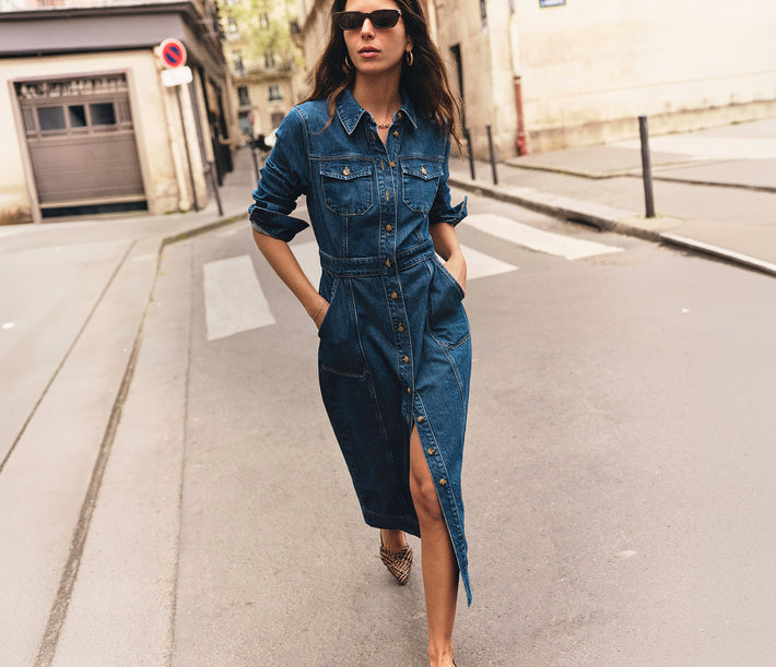 A woman wearing a dark blue denim shirt dress walks down a city street. The dress has a button-front closure, chest pockets, and a high slit on one side.
