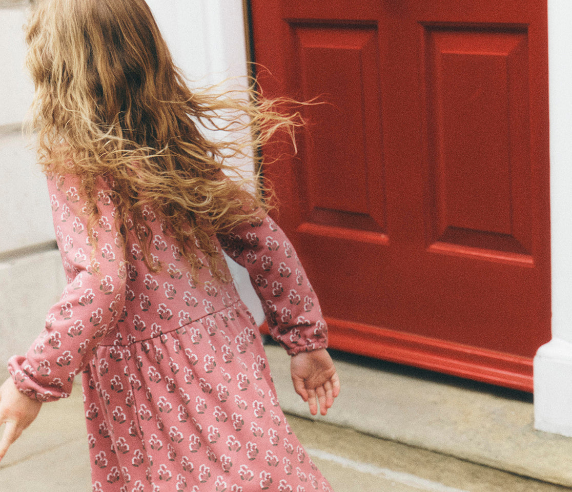 A young girl with long wavy light brown hair is seen from behind, wearing a pink long-sleeve dress with a small white floral pattern, standing in front of a bright red door.