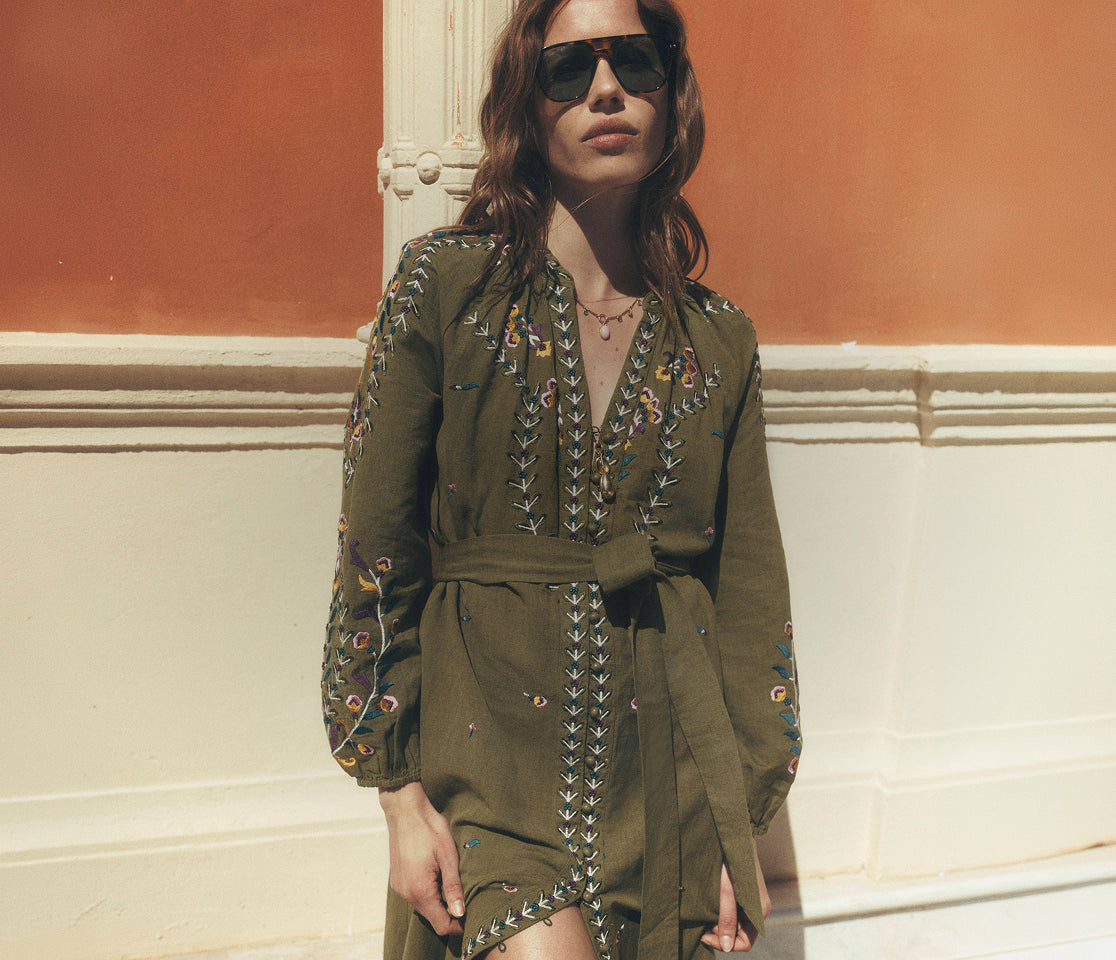 Woman wearing an olive green dress with intricate patterns and sunglasses, standing against a warm-toned wall.