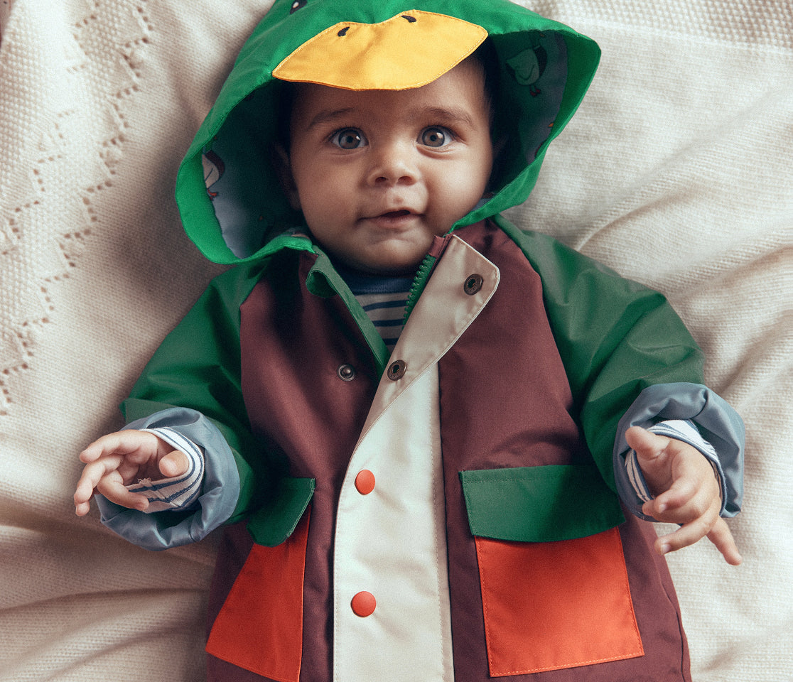 A baby lying on a cream textured blanket, wearing a duck-themed hooded raincoat with green, brown, cream, and orange panels, and a yellow duck face on the hood.
