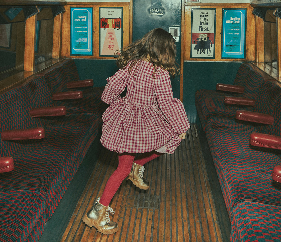 A young girl in a red and white gingham dress, pink tights, and gold boots stands in the aisle of a vintage bus interior, looking back over her shoulder. The bus has patterned red and blue seats with wooden paneling and posters on the walls.