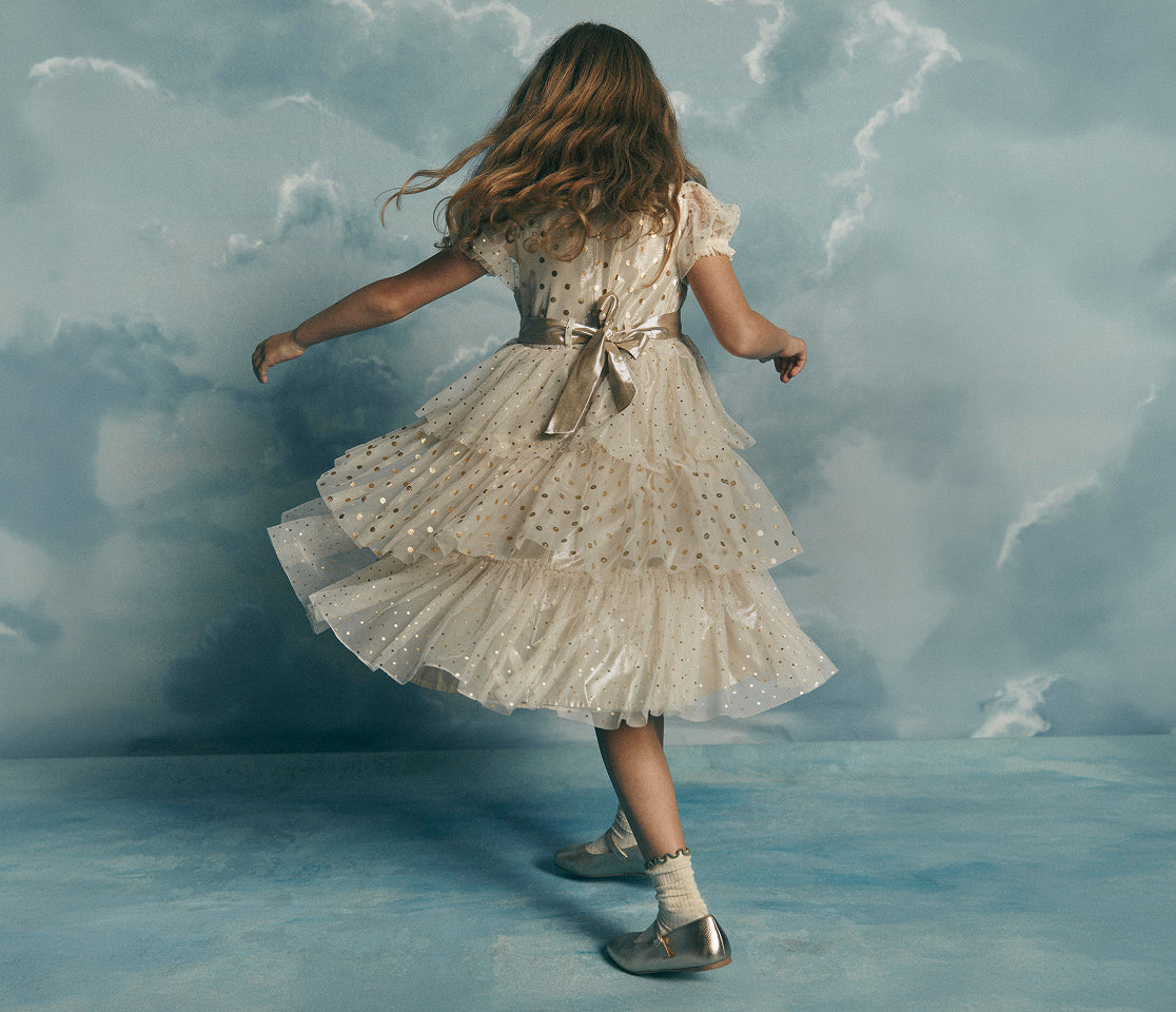 A young girl with long light brown hair is seen from behind, wearing a sparkly cream-colored tiered tulle dress with a bow at the back waist, white socks, and silver shoes, posing against a cloudy blue backdrop.