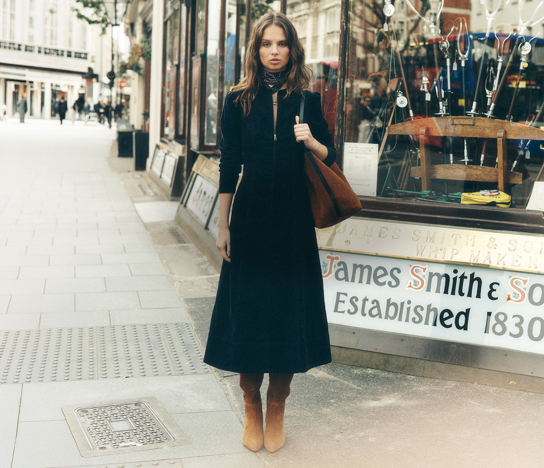 A woman stands on a city sidewalk in front of a shop window, wearing a long black midi coat over a black top and skirt, with tan ankle boots and a brown shoulder bag.