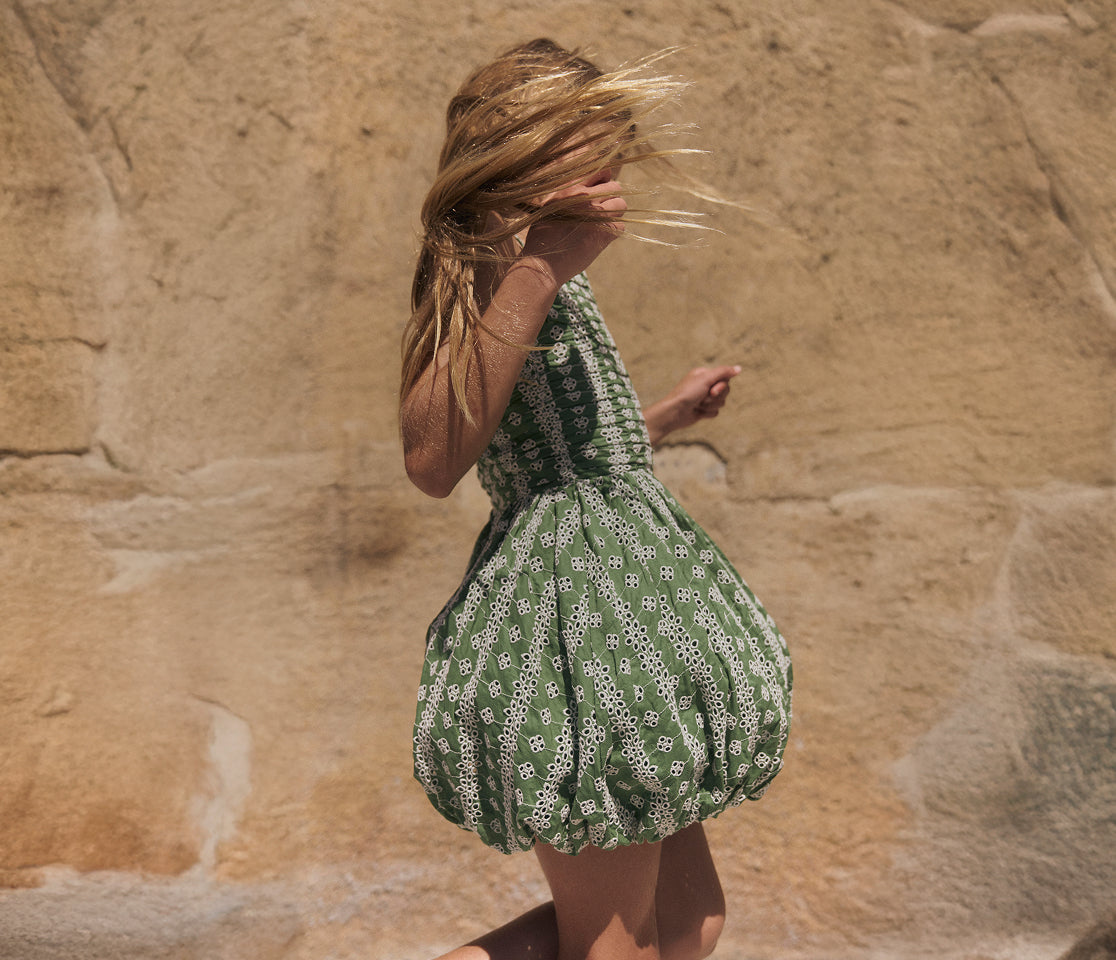 Woman in a green dress with white patterns walking against a beige stone wall.