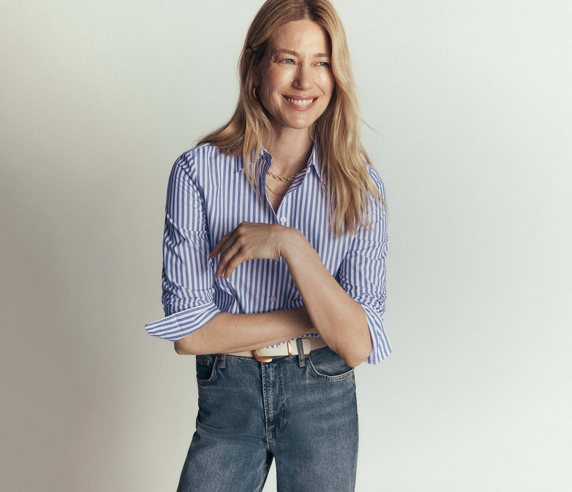 A woman wearing a blue and white vertically striped button-down shirt with rolled-up sleeves and blue jeans, standing against a light gray studio background.