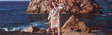 Woman in a colorful dress standing on a rocky coastline with ocean waves.