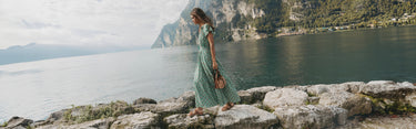 Woman in a green dress standing on rocks by a lake with mountains in the background