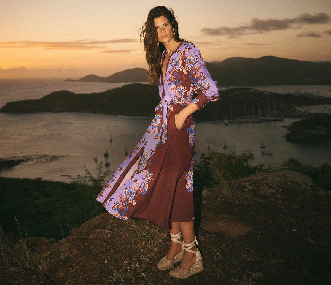 Woman in a floral dress standing on a rocky outcrop with a scenic view of water and mountains.
