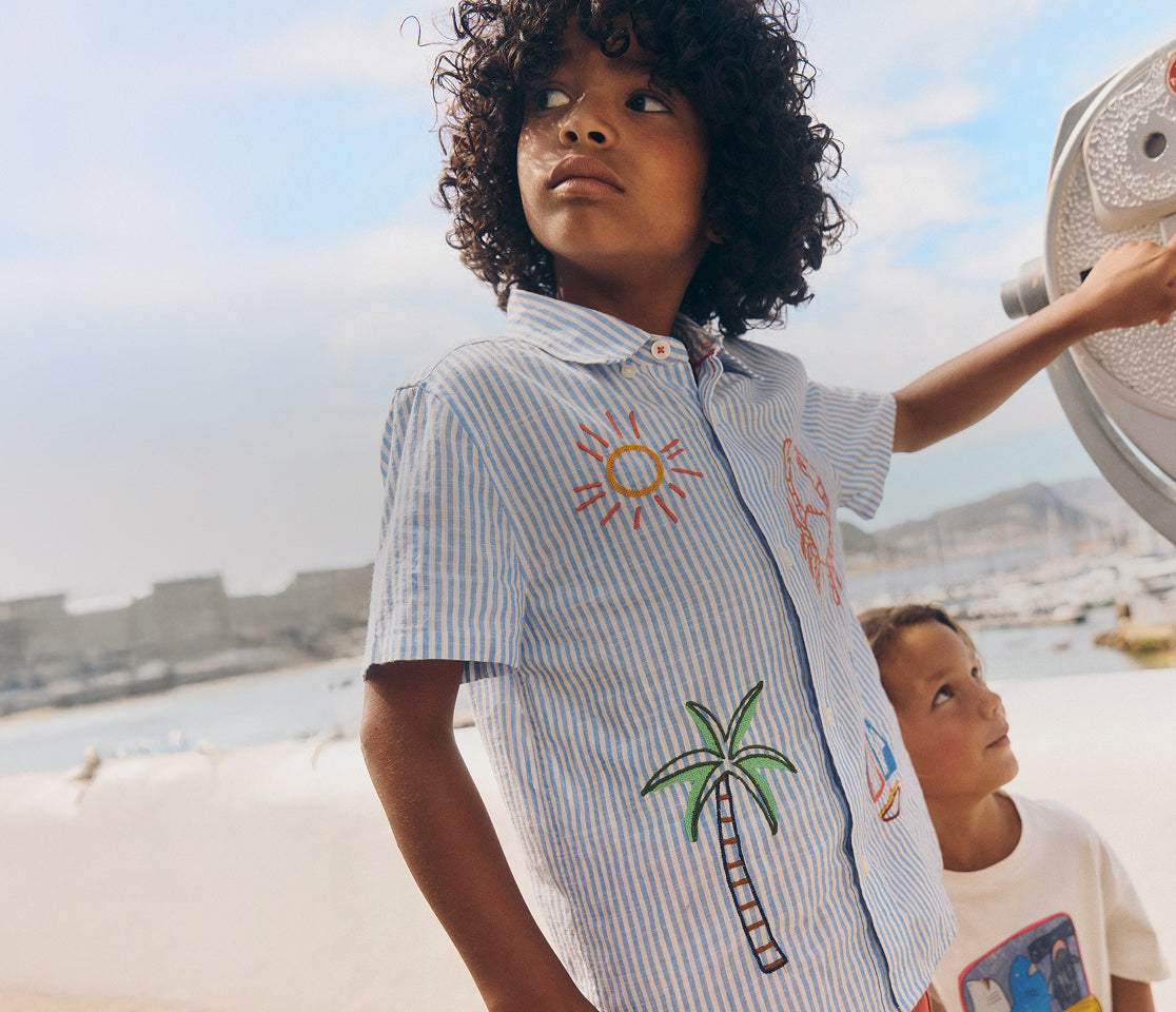 Child wearing a shirt with sun and palm tree designs by a waterfront.