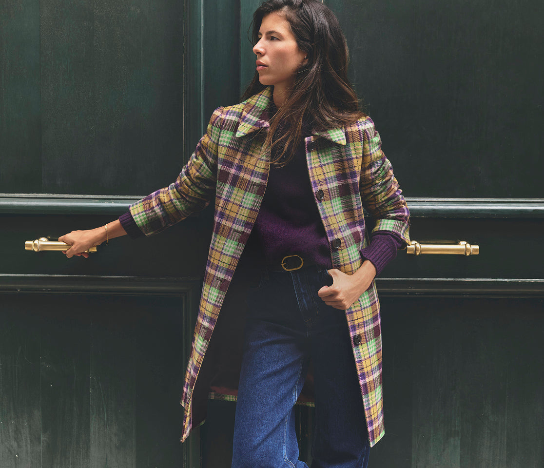 Close-up of a person wearing a purple knit sweater layered under a colourful plaid coat in shades of yellow, green and purple. The outfit is paired with dark denim jeans and a black belt with a gold oval buckle, with both hands tucked into the front pockets.