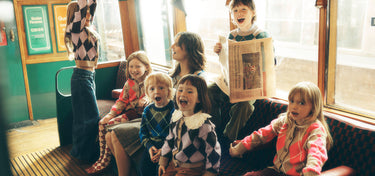 A group of children and a woman sitting on a vintage tram or trolley bus, with one child holding a newspaper. The scene is shot in warm, golden-hour light with a soft focus, highlighting the children's clothing.