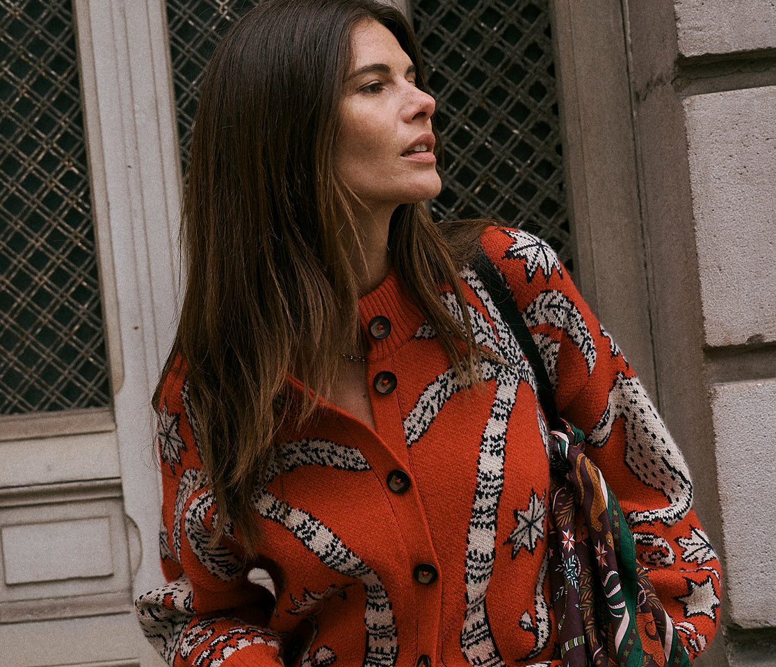 A woman wearing a vibrant orange knit cardigan with a white and black botanical pattern, standing outdoors against a stone wall.