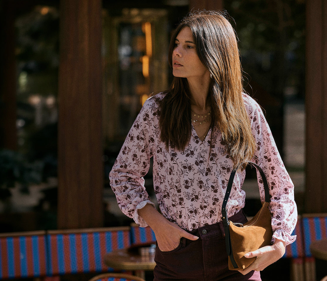 A woman wearing a pink floral long-sleeve blouse and dark brown pants, holding a brown leather shoulder bag in an outdoor patio setting.