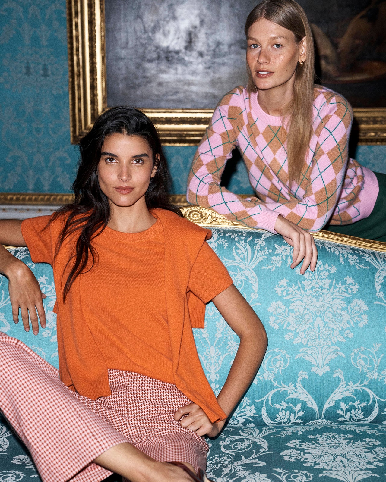 Two models posing on a turquoise damask sofa. The model in the foreground wears an orange short-sleeve top and pink gingham pants.
