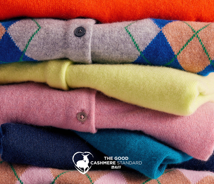 A stack of folded cashmere sweaters in various colors and argyle patterns, including orange, gray, blue, yellow, pink, teal, and navy, with a logo for 'The Good Cashmere Standard' at the bottom.