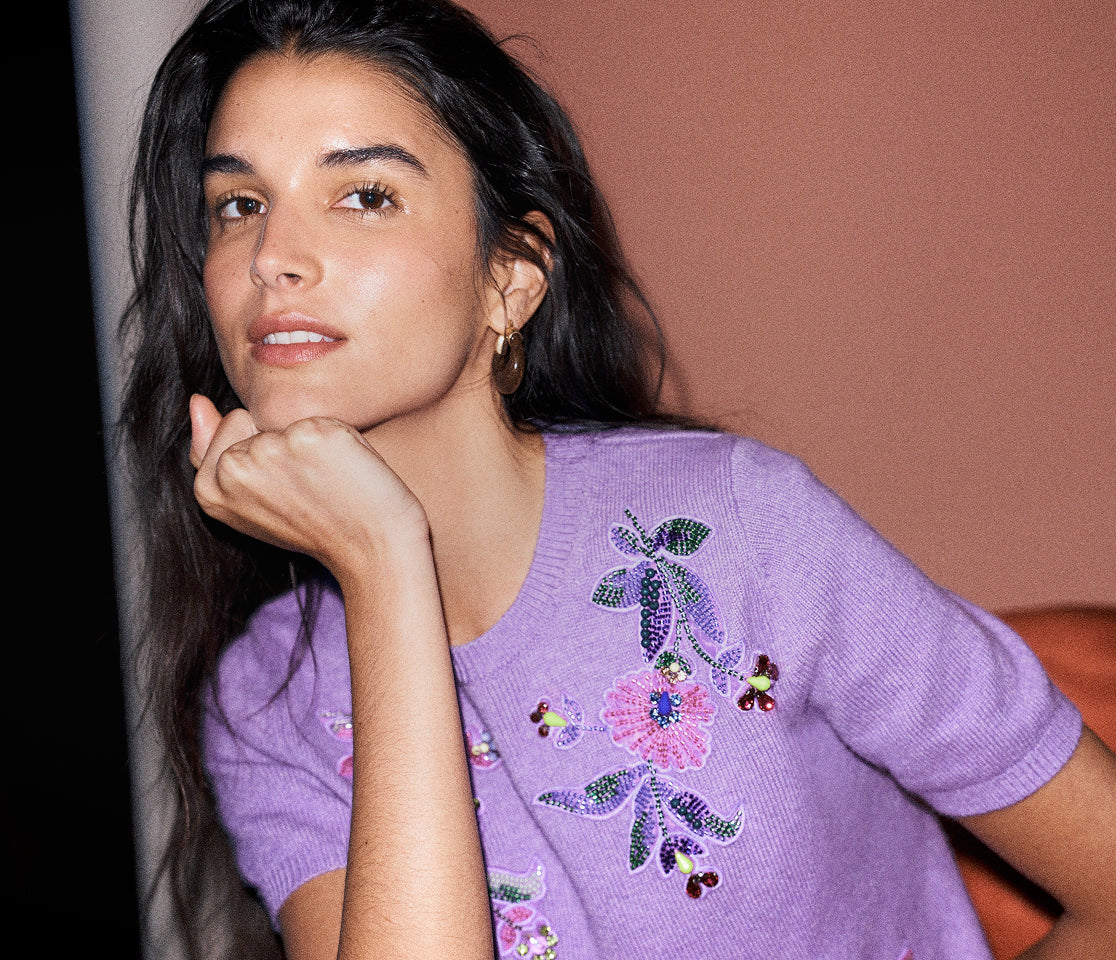 A woman with long dark hair rests her chin on her hand, wearing a lavender short-sleeve knit sweater with colorful floral and butterfly embroidery. She is posed against a warm, solid terracotta-colored background.