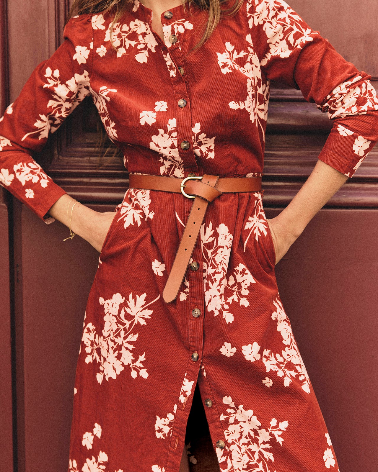 A woman wearing a rust red long-sleeve button-front dress with a white floral print, styled with a brown leather belt, standing against a dark brown wooden door.