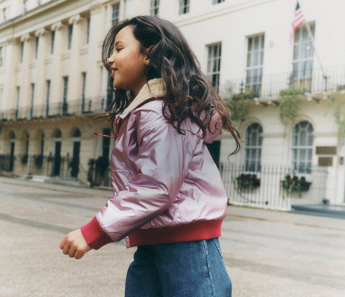 A young girl wearing a shiny light pink bomber jacket with a beige collar and red ribbed trim, walking on a city street with a classic European-style building in the background.