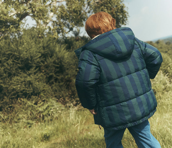 Child photographed from behind walking through a grassy, fern-filled landscape, wearing a hooded puffer jacket with wide navy and dark green vertical stripes, paired with blue jeans.