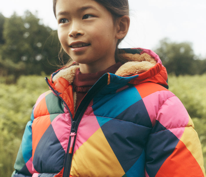 Close-up of a child outdoors wearing a colourful geometric puffer jacket in pink, orange, blue, green and yellow. The hood is lined with soft cream fleece, with a blurred green countryside landscape in the background.
