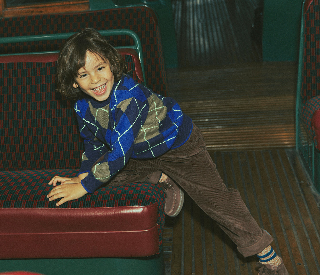 A young boy smiling while sitting on a vintage red and green patterned bus seat, wearing a blue, brown, and gray argyle sweater and brown pants.