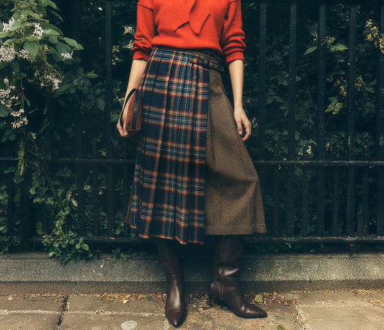 A person standing outdoors in front of a black metal fence and greenery, wearing a bright red long-sleeve top, a pleated plaid skirt with a brown side panel, and dark brown knee-high boots.