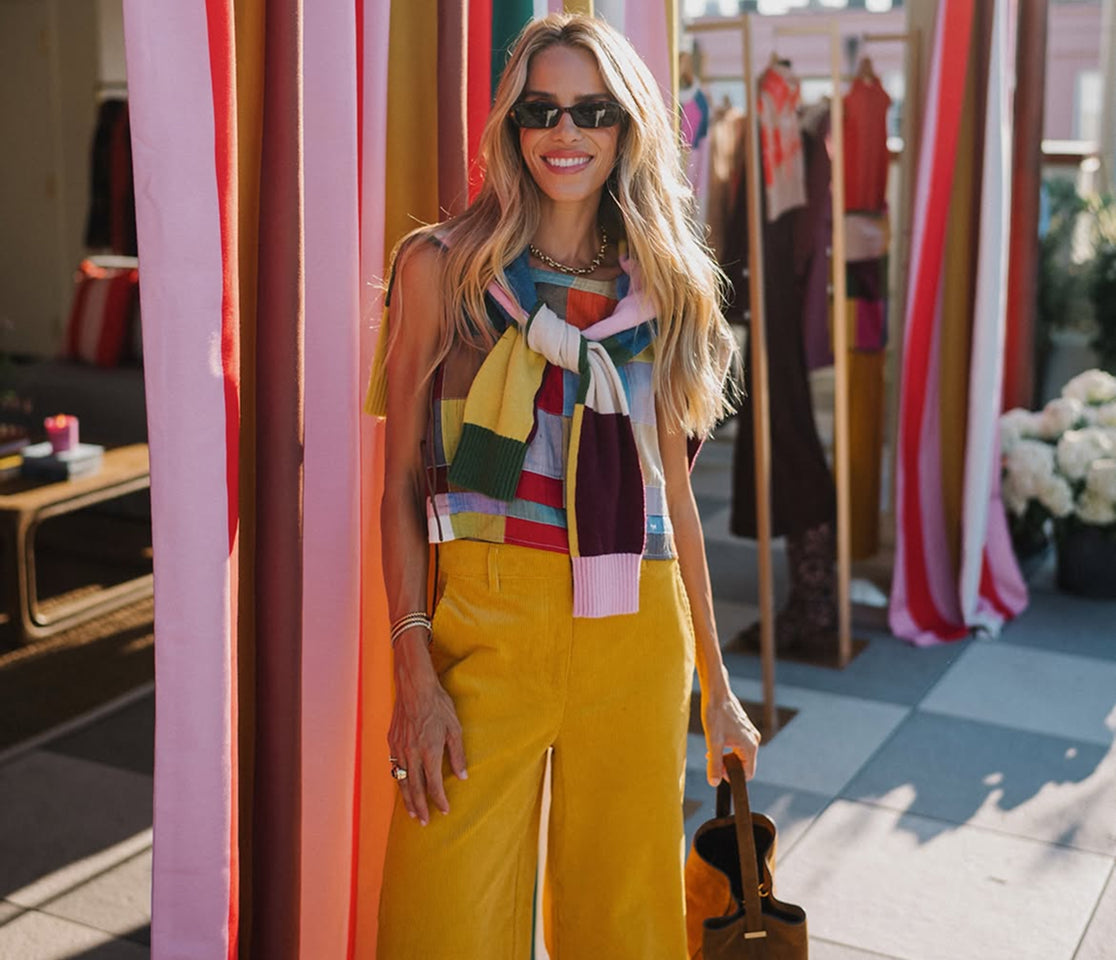 Women wearing sunglasses, mustard yellow trousers and a colourful striped top with a coordinating knit draped over the shoulders, standing among vibrant fabric panels.