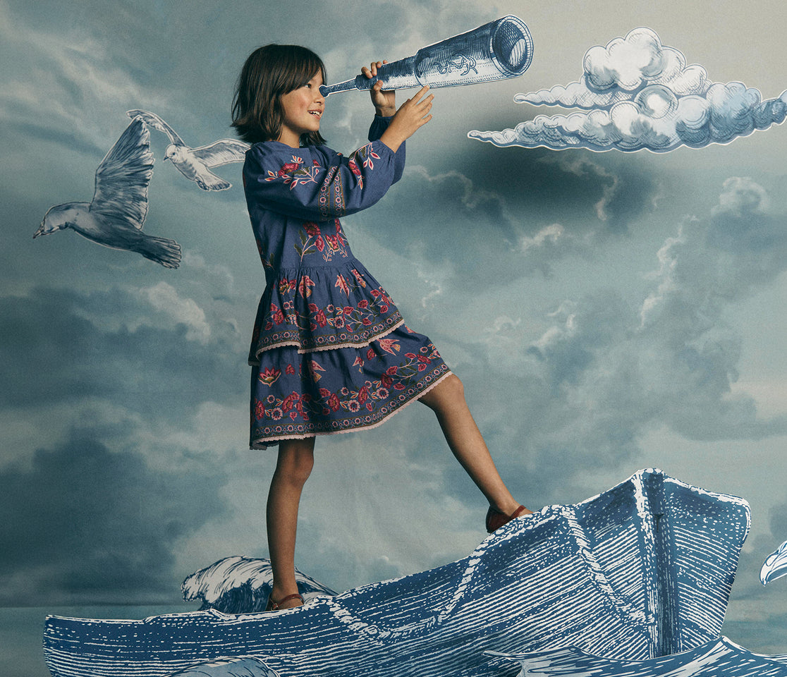 A young girl in a blue floral dress stands on a stylized boat against a cloudy sky background, looking through a bottle-shaped telescope. A seagull flies beside her.