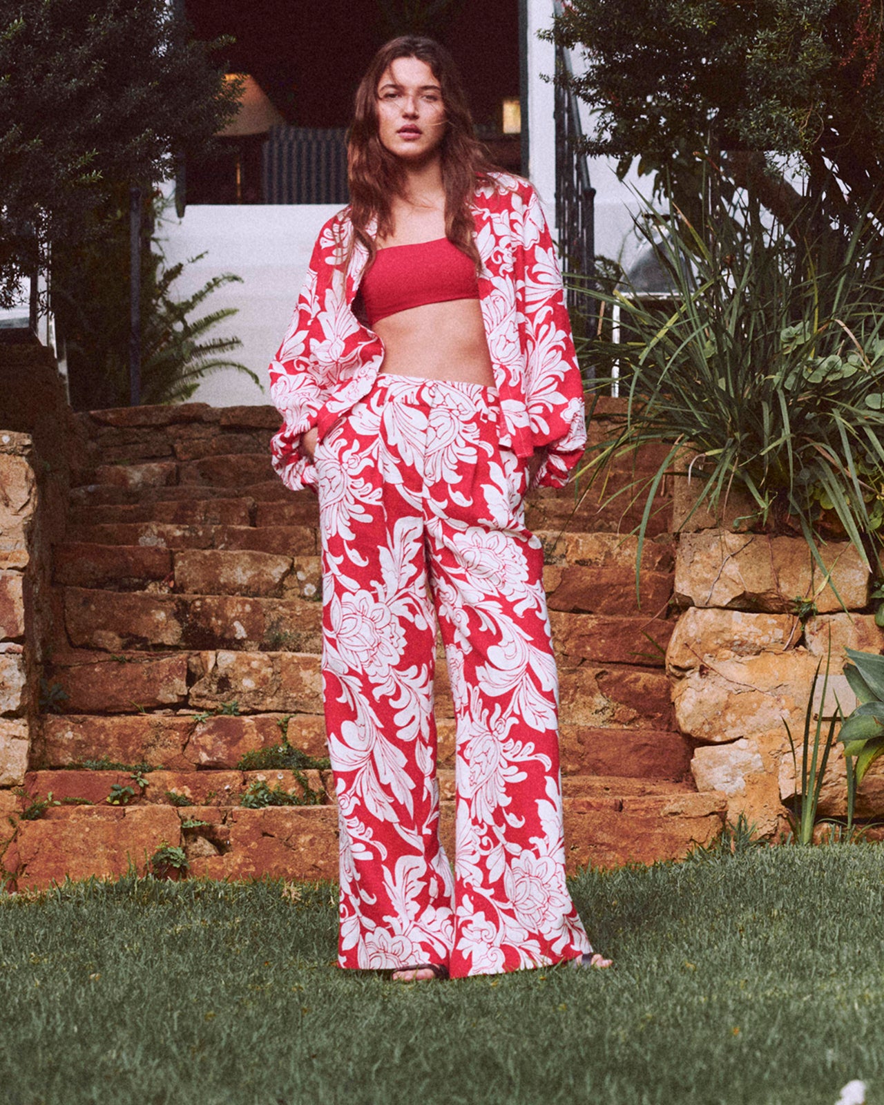 Woman wearing a red and white floral outfit standing outdoors with stone wall and plants in the background