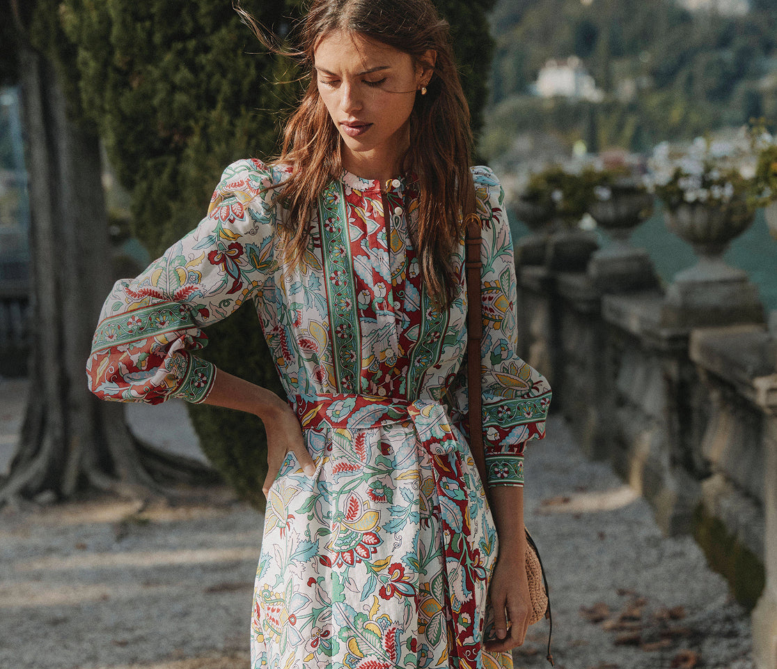 Woman in a colorful floral dress standing outdoors with greenery and stone steps in the background